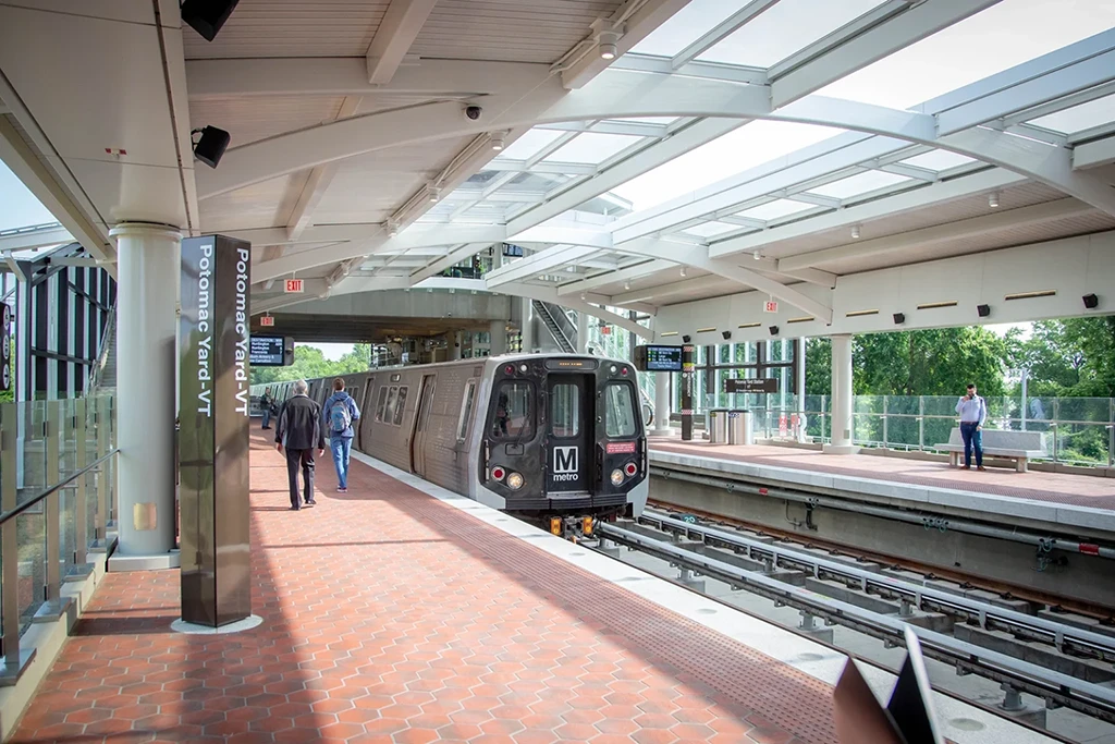 A Washington DC Metro train arrives at the Potomac Yard station, featuring modern architecture with a bright, open design, and passengers walking along the platform under a glass roof. The scene highlights the system's anniversary and commitment to improved transit.