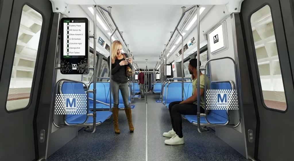 A modern Metro train interior with blue seats, featuring a woman using her phone and a seated man. Metro signage is visible, celebrating the 50th anniversary of the Washington DC Metro system. The environment is bright and clean, showcasing the design of the updated train.