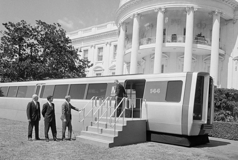 A vintage black and white photograph depicting a futuristic Metro train model beside the White House, with four men in suits, one greeting another stepping off the train. The scene marks a significant moment in the history of the Washington DC Metro system during its 50th anniversary celebrations.