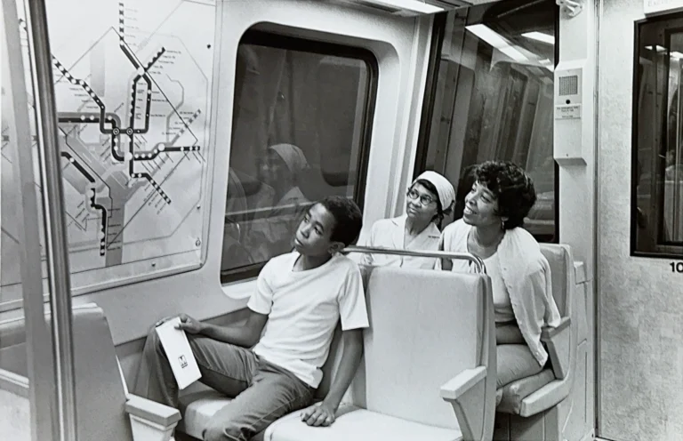 Three passengers inside a Washington DC Metro train, smiling and observing a map of the Metro system. The interior is clean and modern, with light-colored seats. This black-and-white photo reflects the community's engagement with public transportation as part of the Metro's 50th anniversary celebration.