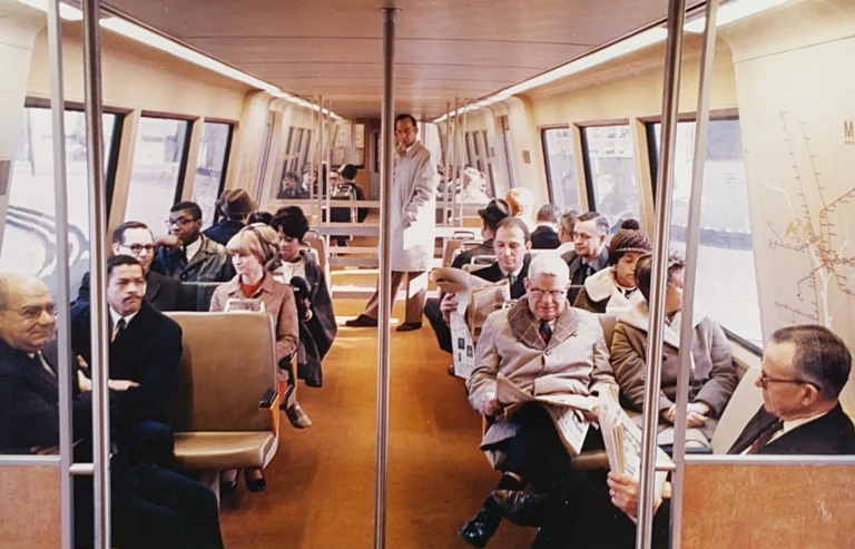 The interior of a 1970s Washington DC Metro train, featuring seated passengers reading newspapers, dressed in period attire. A conductor stands at the end of the car. The warm-toned decor reflects the design of the era, and a map of the Metro system is visible on the wall, celebrating the system's 50th anniversary.