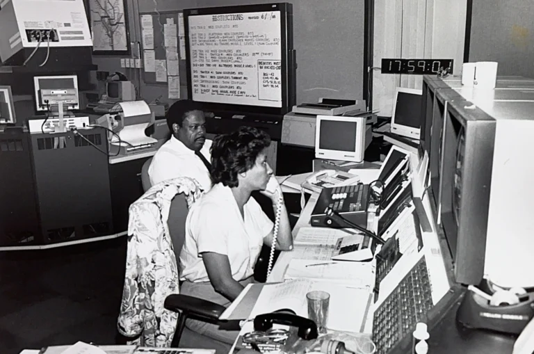 Two operators are working in a control center, one on the phone while the other monitors multiple computer screens. In the background, a large clock shows the time as 17:59, and a whiteboard with handwritten notes is mounted on the wall, reflecting a busy environment related to the Washington DC Metro system.