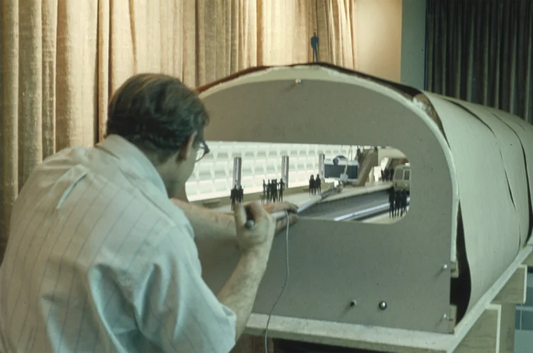A person working on a model of a Metro train, focusing on the interior design and layout, with figures representing passengers and a detailed model environment in a workshop setting. The background features draped curtains, suggesting a staged or presentation area.
