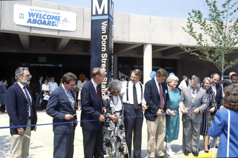 A group of dignitaries and officials gather outside the Van Dorn Street Metro Station, holding scissors and preparing to cut a ribbon. The scene features banners celebrating the 50th anniversary of the Metro system, with visible landscaping and a modern building in the background.