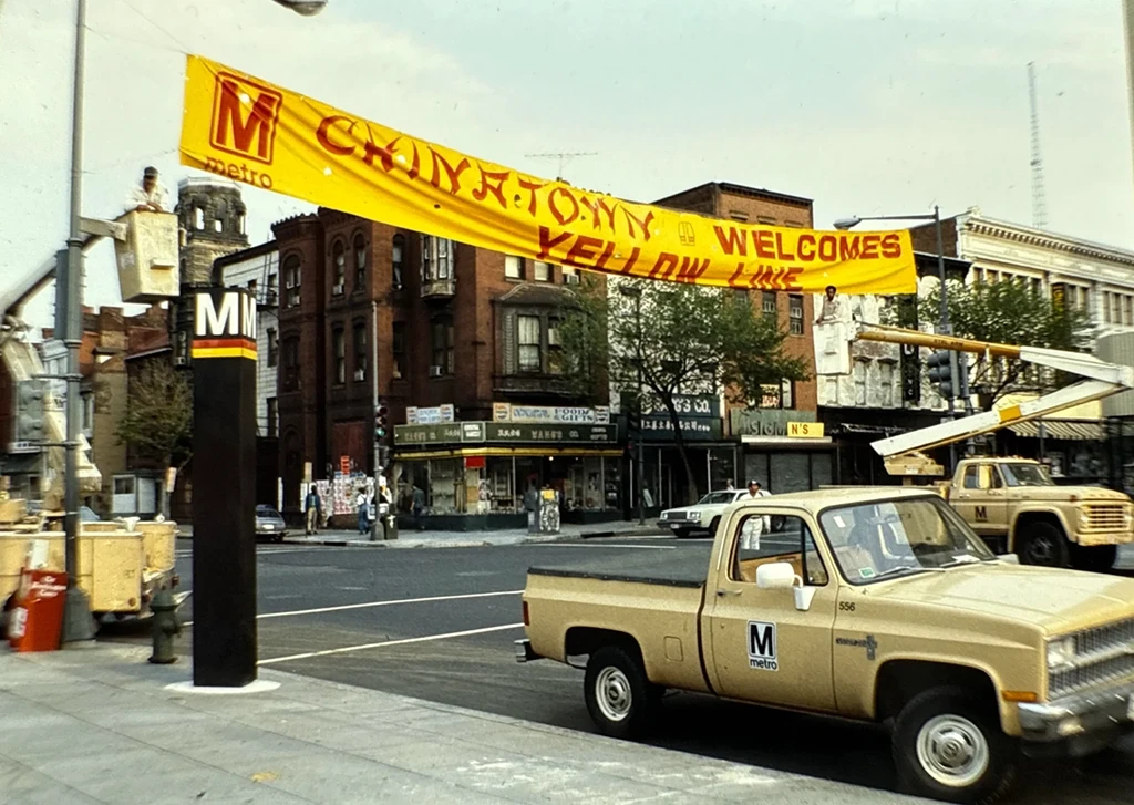 A vibrant welcome banner reads "Metro Welcomes Yellow Line" across a street, with trucks and construction equipment nearby. Historical buildings and shops are seen in the background, capturing the atmosphere of a celebration for the 50th anniversary of the Metro system in Washington DC.