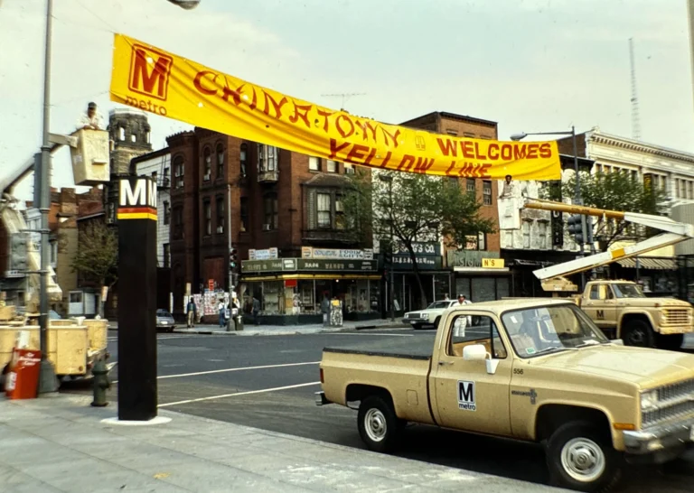 A large yellow banner reading "Chinatown Welcomes Yellow Line" spans across a street, with a vintage pickup truck parked nearby. The scene features the Metro logo and construction equipment, highlighting preparations for the Yellow Line opening in Washington, D.C. storefronts and a busy street are visible in the background.
