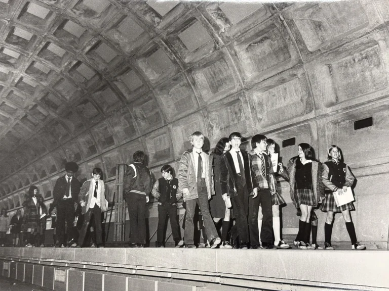A group of students dressed in 1970s fashion, including skirts and suits, stands on a Metro platform in a vacant subway station. The platform features a distinctive textured wall, symbolizing the era of the Metro system. The photograph captures a moment in time, celebrating the history of the Washington DC Metro.