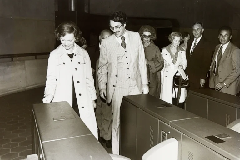 A group of people, including a woman in a light trench coat and a man in a striped suit, pass through fare gates in a Metro station. Other attendees, dressed in 1970s fashion, look on, adding to the historic atmosphere of the event.