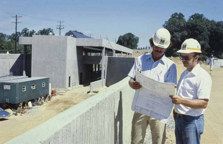 Two construction workers in hard hats examine blueprints at a transit site, with a partially completed station in the background. The setting includes machinery, a trailer, and trees, reflecting the development of the Washington DC Metro system during its construction phase.
