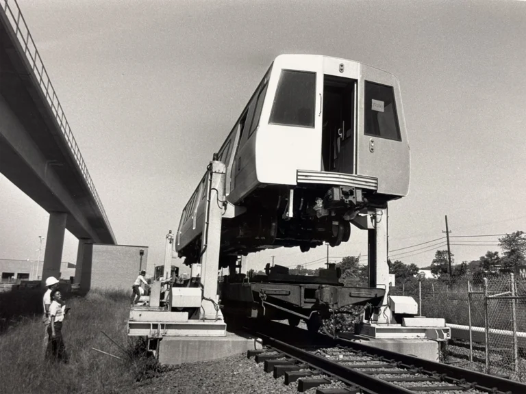 A vintage black and white photo showing a Metro train car being lifted on a platform near a railway track, with workers in hard hats observing and a bridge in the background, commemorating the 50th anniversary of the Metro system in the Washington DC region.