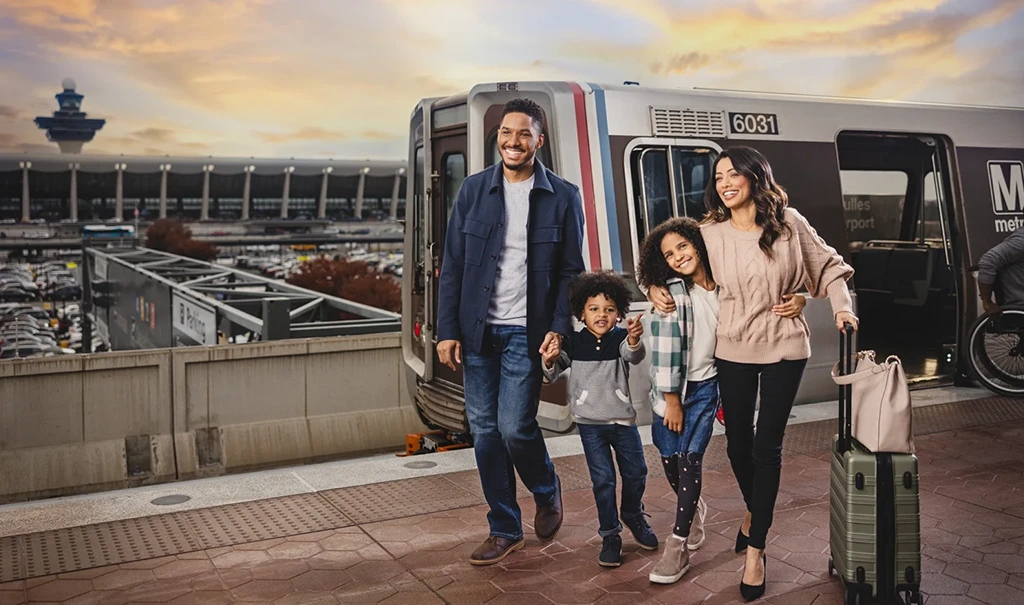 A smiling family of four, including a father, mother, and two children, stands at a Metro station with luggage. They appear happy and excited, with a Metro train in the background. The setting features a well-lit terminal and a parking area, emphasizing the convenience of the Metro system for travel in celebration of its 50th anniversary.