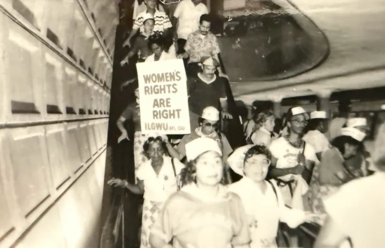 Black and white photo of people descending an escalator in a Metro station, some wearing hats. A person holds a sign reading "WOMEN'S RIGHTS ARE RIGHT," emphasizing a rally or protest related to women's rights, reflecting social movements relevant to the Metro's 50th anniversary.