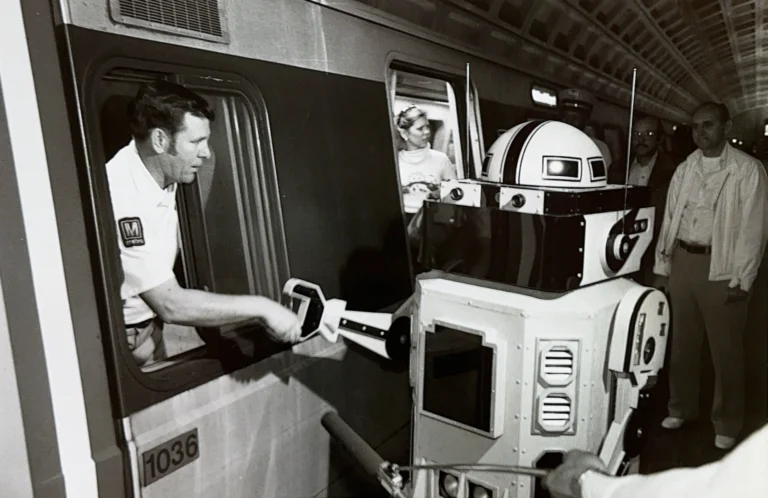 A Metro train operator hands a robotic character a stylized device at a Washington DC Metro station, surrounded by onlookers, celebrating the 50th anniversary of the Metro system. The scene highlights the intersection of technology and public transit in a historical context.