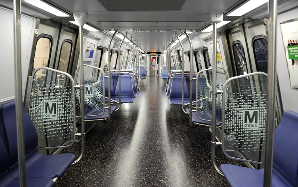 Interior of a Washington DC Metro train showing empty rows of blue seats, metallic poles, and floor-to-ceiling windows, highlighting a clean, modern design. The train is illuminated with overhead lights, and the doors are closed, illustrating a typical transit environment for the Metro system's 50th anniversary.