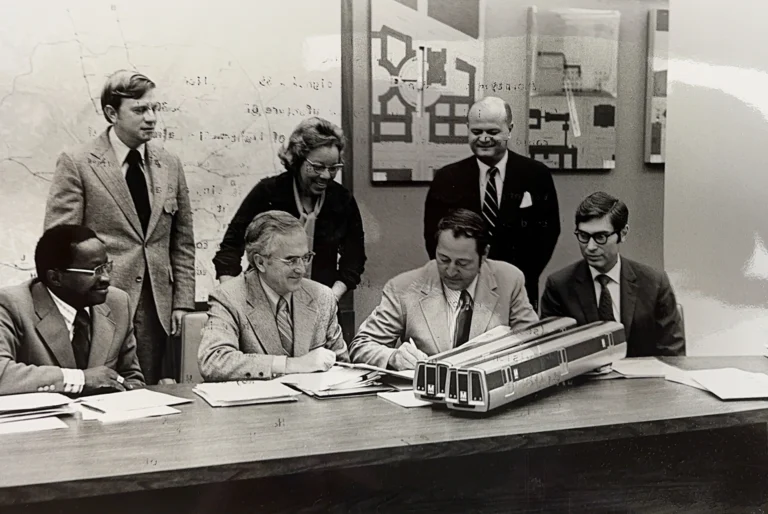 A group of six people gathered around a conference table, examining documents and a model train, likely discussing plans for the Washington DC Metro system, with maps displayed on the wall in the background. The scene captures a pivotal planning moment for the Metro's development.