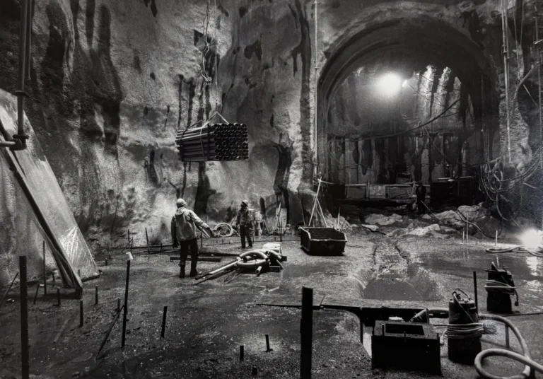 Construction workers operate in a large underground tunnel during the building of the Washington DC Metro system, with equipment and scaffolding visible amidst the rocky walls and wet floor, highlighting the engineering efforts of the time.