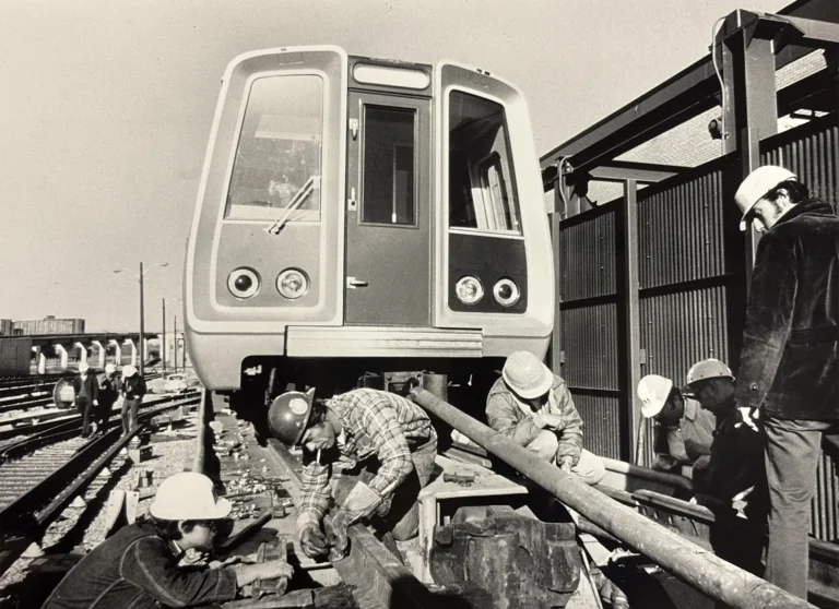 A black and white photo showing workers in hard hats assembling tracks for the Washington DC Metro system, with a train in the background. The scene captures the construction phase, highlighting teamwork and infrastructure development for the Metro's launch.
