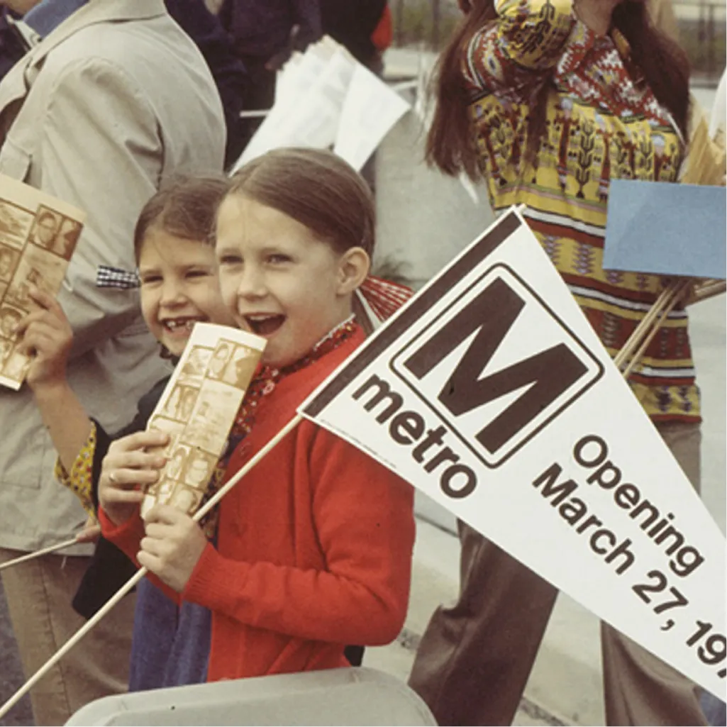 Two young girls smile while holding Metro flags and brochures, celebrating the opening of the Washington DC Metro system on March 27, 1976. They are part of a crowd, showcasing excitement and nostalgia for the 50th anniversary of the Metro. The background suggests a festive atmosphere.