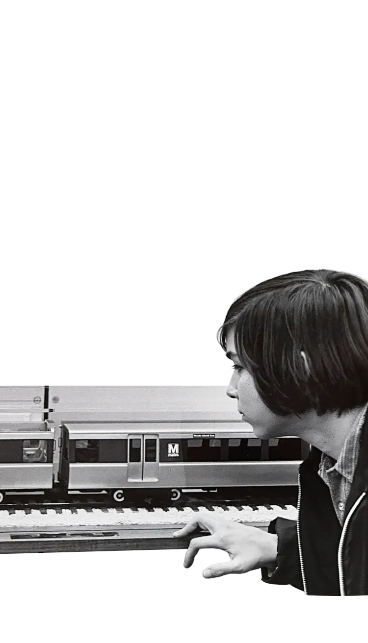 A young boy observes a detailed model of a Washington DC Metro train, featuring the iconic red and silver colors with the Metro logo. The scene captures his focused expression as he leans in, creating a nostalgic connection to the Metro system's history.