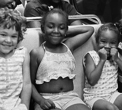 Three smiling young girls sit together on a subway train, showcasing a moment of joy and friendship. The girl in the center wears a cropped top, while the others display playful expressions in casual clothing, reflecting the vibrant community atmosphere of the Metro.