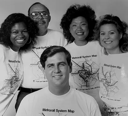 A group of five people poses together, all wearing white t-shirts featuring a Metrorail system map. The individuals include three women and two men, showcasing a diverse group. Their expressions range from smiles to laughter, reflecting a celebratory mood, in honor of the Washington DC Metro system's 50th anniversary.