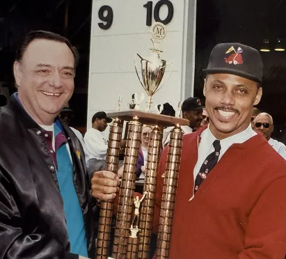 Two men smile while holding a large trophy, celebrating an achievement. The backdrop features the number 9 and 10, hinting at a Metro station. The event seems festive, likely in honor of the Metro system's 50th anniversary in Washington, DC.