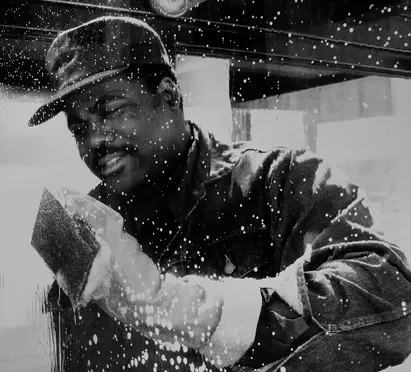 A smiling worker in a cap and gloves cleans a large glass window, creating a sparkling effect with soap suds, highlighting the dedication of staff involved in maintaining the Metro system.