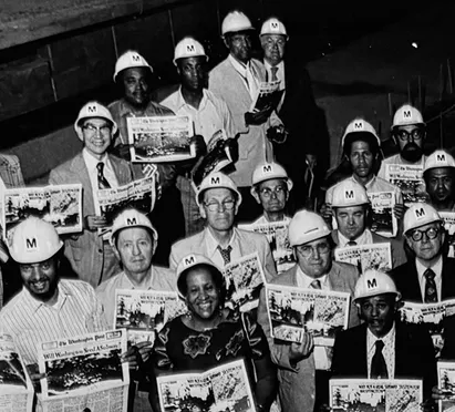 A group of diverse individuals wearing white hard hats with "M" logos, holding newspapers featuring the Washington Metro's history, celebrating the 50th anniversary of the Metro system in Washington, DC. The photo captures a moment of pride and accomplishment in the development of public transit.