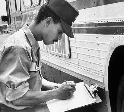 A man in a uniform and cap writes on a clipboard while standing next to a Metro bus, capturing a moment from the early days of the Washington DC Metro system.