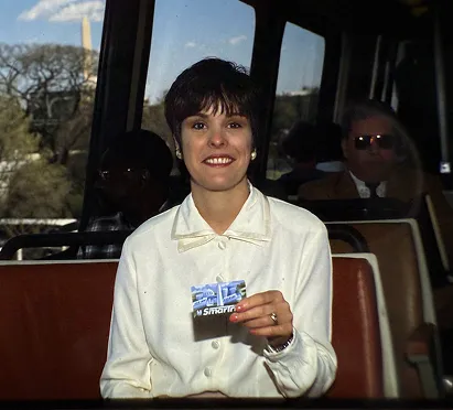 A woman in a white shirt smiles while holding a Metro card on a train, with blurred passengers in the background and a view of trees outside the window. This scene reflects the history and usage of the Washington DC Metro system, celebrating its 50th anniversary.