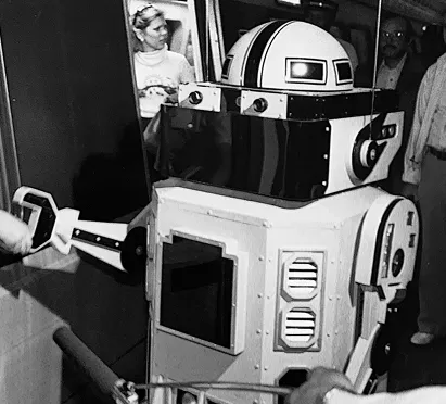 A retro robot character interacts with a crowd in a Washington DC Metro station, celebrating the 50th anniversary of the Metro system. The robot features a distinctive design with a large head and metallic body, while people watch curiously from the surrounding area.