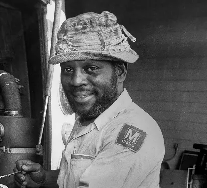 A smiling Metro worker in a light-colored uniform and a worn cap, standing next to equipment, embodies the spirit of service and dedication during the 50th anniversary of the Washington DC Metro system.