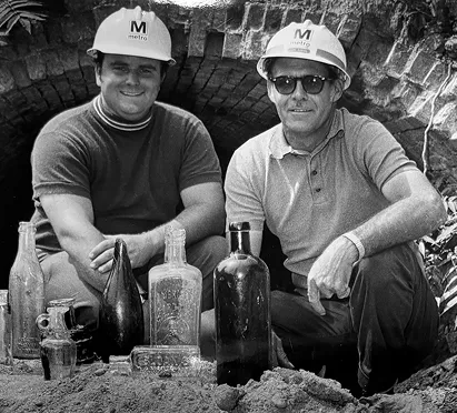 Two men wearing white construction helmets with the Metro logo pose next to a collection of historical bottles. They are kneeling in front of a brick tunnel, showcasing remnants from the past during the excavation for the Washington DC Metro system.