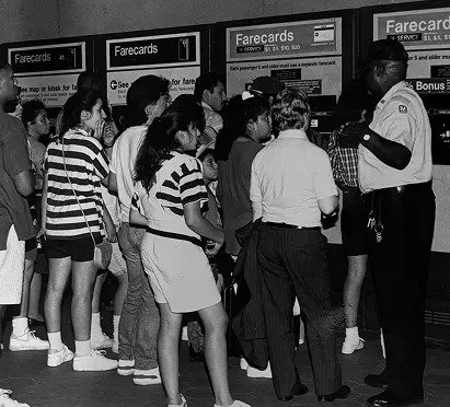 A crowd of diverse people, including children and adults, stands in front of farecard vending machines at a Metro station, listening to an employee in uniform explaining the fare system. The scene captures the busyness and excitement of public transit, highlighting the community engagement during the Metro's 50th anniversary celebrations.