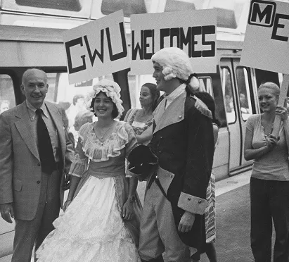 A group celebrating the 50th anniversary of the Metro system, dressed in period costumes, is holding signs that read "GWU WELCOMES." The scene features a man in a suit and two people in 18th-century attire, with a Metro train in the background. The atmosphere is festive and historical, highlighting community support for the event.