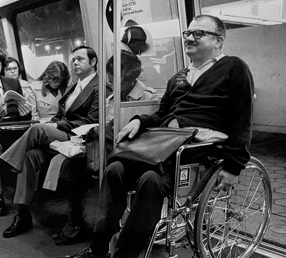 A black and white photograph of a man in a wheelchair on a Metro train, smiling while surrounded by seated passengers. The setting reflects the atmosphere of public transportation, highlighting accessibility and the diversity of riders.
