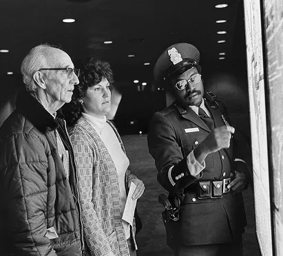Two people, an elderly man and a woman, observe a display with a police officer pointing at it. The setting appears to be a Metro station. The black and white photo evokes a sense of nostalgia, reflecting on the history and community connections of the Metro system.