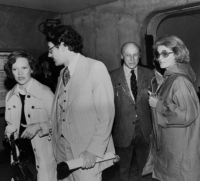 A group of four individuals in a subway station, dressed in 1970s fashion. Two women, one in a light coat and the other wearing glasses, are accompanied by two men in suits. They appear to be engaged in conversation, reflecting a moment from the Metro system's early years. The setting features a simple subway decor.