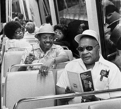 Two men sit on a Metro train, one reading a booklet while the other smiles and holds a drink. Behind them, passengers enjoy the ride, showcasing a mix of hairstyles and 1970s fashion. The black-and-white image captures a moment of camaraderie and relaxation on the Washington DC Metro, emphasizing the public transport culture.