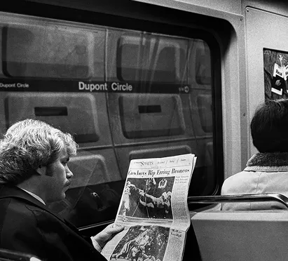 A passenger reading a newspaper on the Metro with a focus on Dupont Circle station visible through the window, capturing a moment of daily commuting. The scene reflects the long history and significance of the Washington DC Metro system as it celebrates its 50th anniversary.