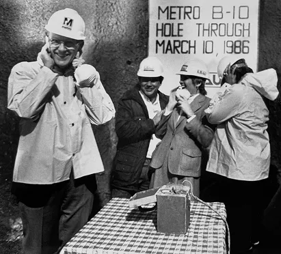 A group of people in hard hats celebrates the construction of the Metro system with a large sign reading "METRO B-10 HOLE THROUGH MARCH 10, 1986." They are gathered around a table with a cake, enjoying the milestone moment during the metro's early development phase.