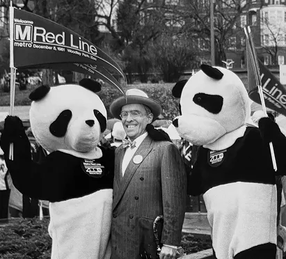 Two people dressed in panda costumes stand beside a man in a suit and bowler hat, smiling as they pose for a photo. A banner welcomes the Metro Red Line, celebrating its anniversary, while a crowd and trees are visible in the background, capturing a festive atmosphere.