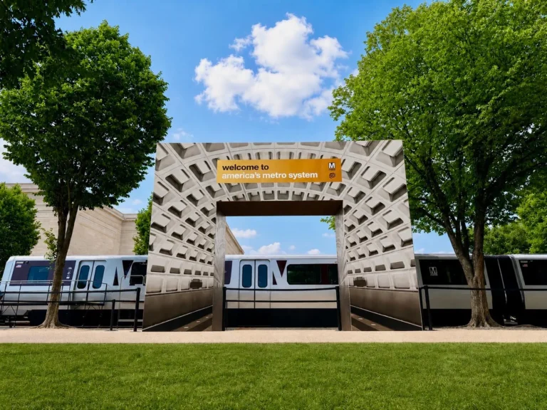 A modern Metro station entrance framed by trees, featuring a large welcoming sign that reads "welcome to America's metro system," with a train visible in the background and a clear blue sky above.