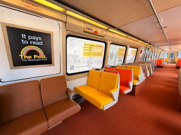 Interior of a Washington DC Metro train, featuring a mix of brown and orange seats. A prominent advertisement on the wall promotes reading The Washington Post, accompanied by various yellow informational posters celebrating the Metro's 50th anniversary. The train interior has a modern design with a clean, bright aesthetic.