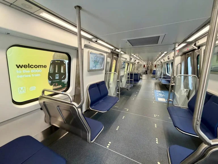 Inside view of a modern Metro train car featuring blue seats and clear signage; a yellow advertisement welcomes passengers to the 8000 series train, celebrating the 50th anniversary of the Washington DC Metro system. The car is well-lit and spacious, with a map displayed on the wall.