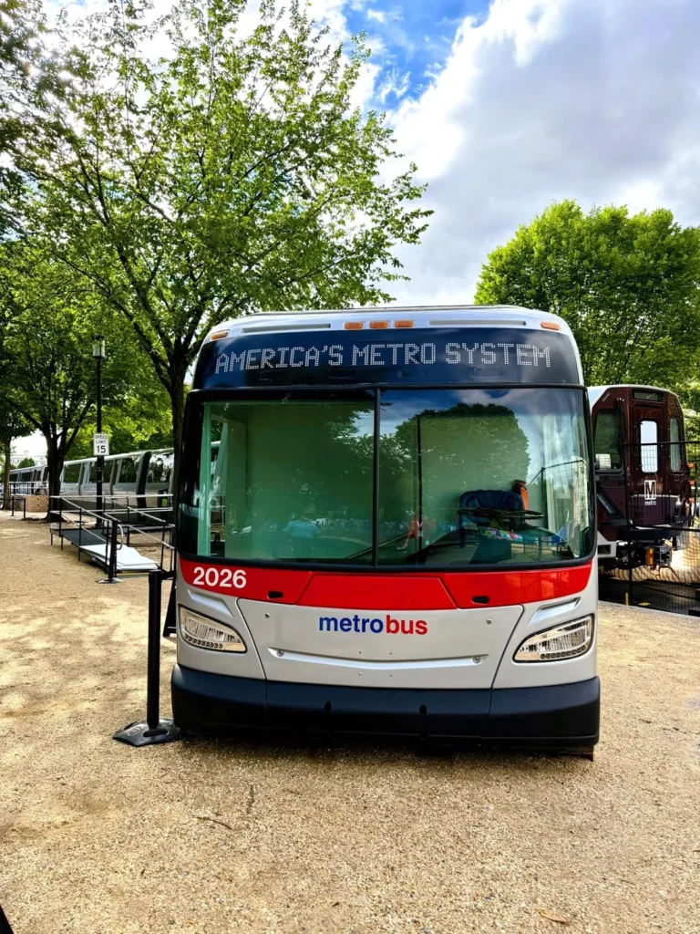 A Metrobus marked "2026" is parked on a gravel path, displaying the text "AMERICA'S METRO SYSTEM" on its front. Lush green trees line the background under a partly cloudy sky, adding to the celebratory atmosphere of the 50th anniversary of the Washington DC Metro system.