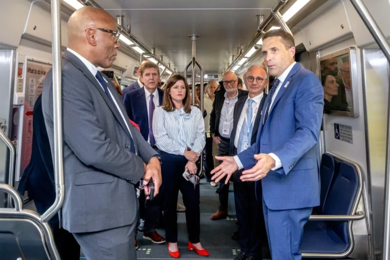 A group of officials in a Metro train cabin, discussing the system’s 50th anniversary. Notable figures include a man in a blue suit gesturing while speaking, surrounded by attendees dressed in business attire, all engaged in conversation amid modern train décor.