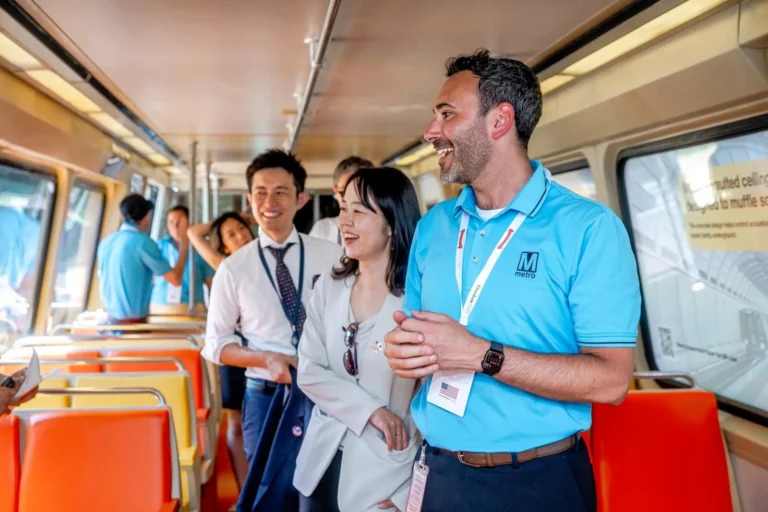 A group of diverse people inside a Washington DC Metro train, smiling and engaging with each other, celebrating the Metro’s 50th anniversary. The man in a blue Metro shirt is laughing while others appear to be enjoying the moment, highlighting a sense of community and joy.