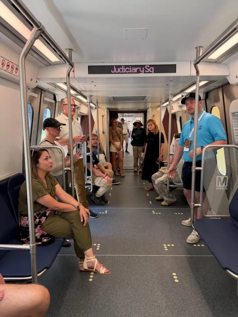 Interior view of a Washington DC Metro train on the 50th anniversary, featuring seated and standing passengers in casual attire, with a prominent sign overhead reading "Judiciary Sq." The atmosphere is relaxed, highlighting the everyday use of the Metro system.