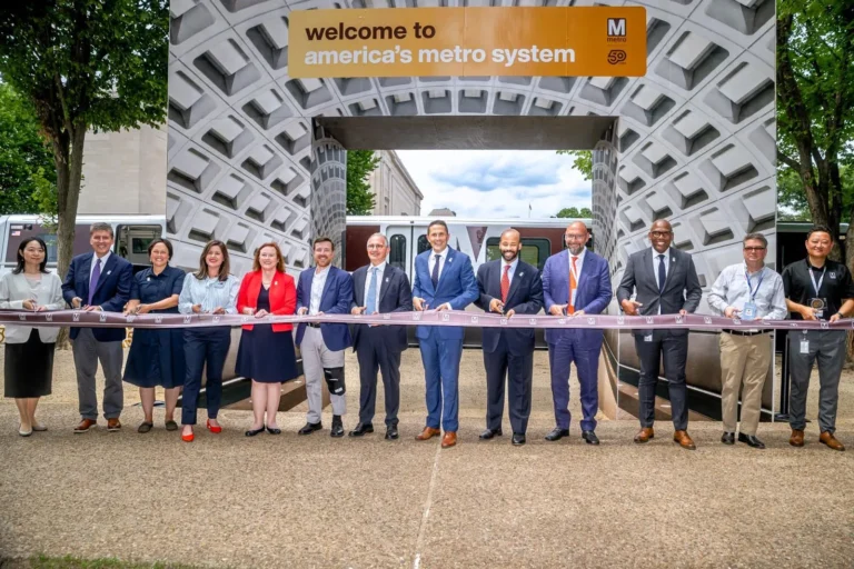 A diverse group of officials and community members gathers to cut a ribbon at a ceremony celebrating the 50th anniversary of the Washington DC Metro system, positioned under an arch that reads "welcome to america’s metro system." The scene features smiles, formal attire, and a backdrop of trees and sky.